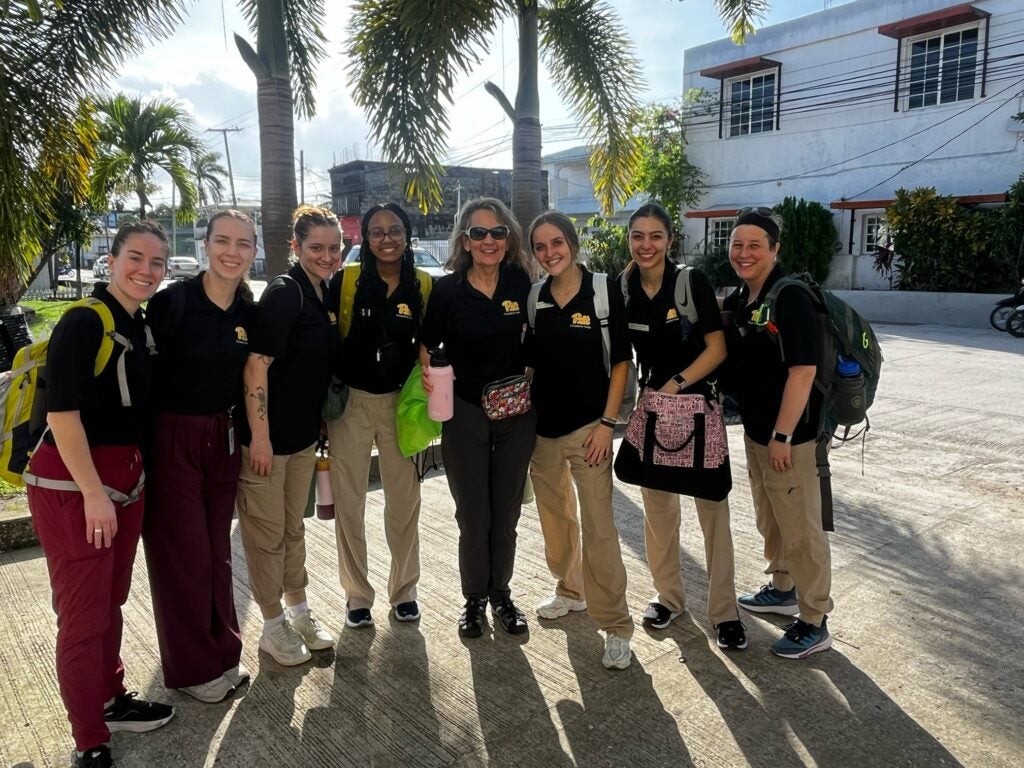 A group of eight women wearing dark Pitt shirts, khaki or dark pants, gym shoes and backpacks while standing together.