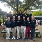A group of 11 women wearing dark Pitt shirts and light and dark pants gathered together in front of a bench.