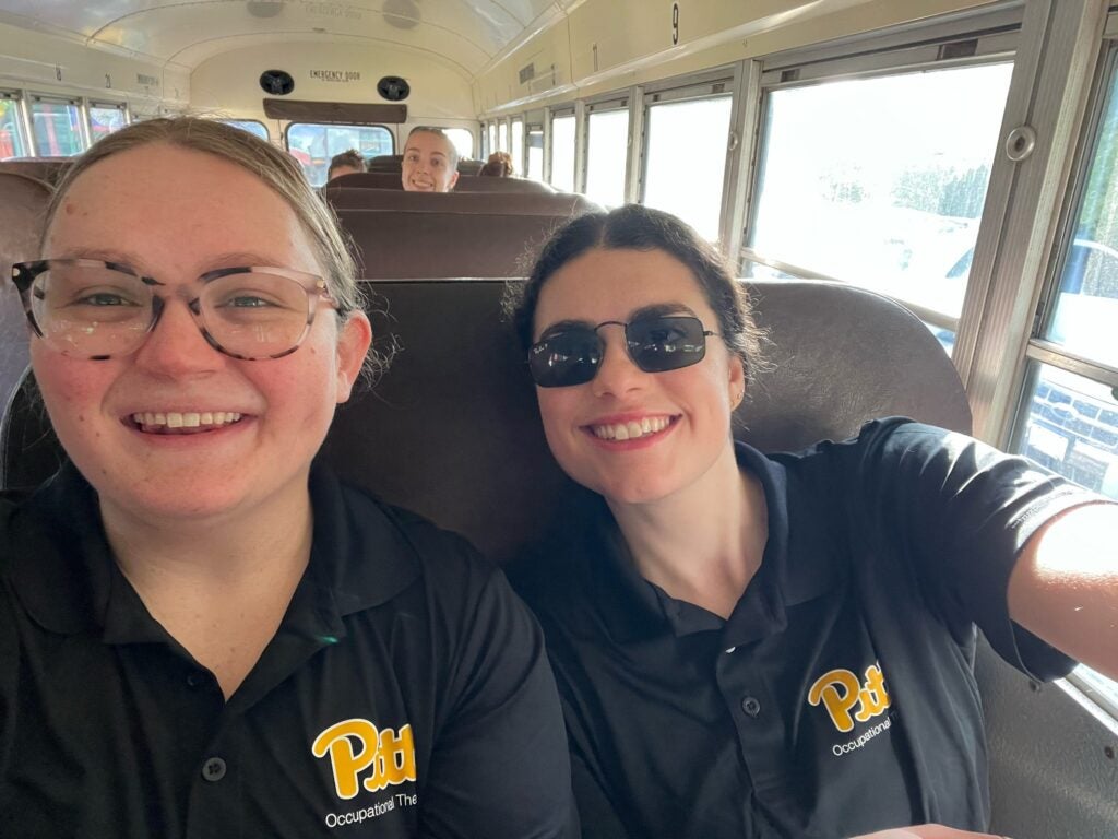 A woman with blonde hair pulled back wearing pink and brown glasses and a black Pitt Occupational Therapy polo shirt sitting in a seat next to a woman with dark hair pulled back who is wearing dark sunglasses and a black Pitt Occupational Therapy polo shirt.