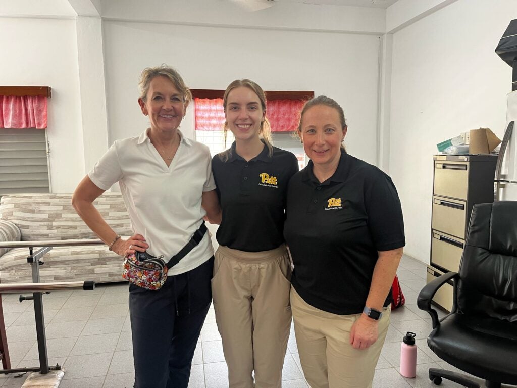 A woman with blonde hair wearing a white collared shirt and dark pants, and two women with dark blonde hair wearing black polo shirts and khaki pants all standing together in a clinical setting.