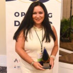 Woman with long dark hair wearing a white dress with white patterned detailing, a pearl necklace and a silver watch while holding a teardrop-shaped award.