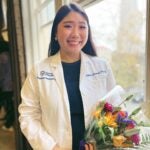 Woman with dark hair wearing a white lab coat over a dark dress and holding flowers.