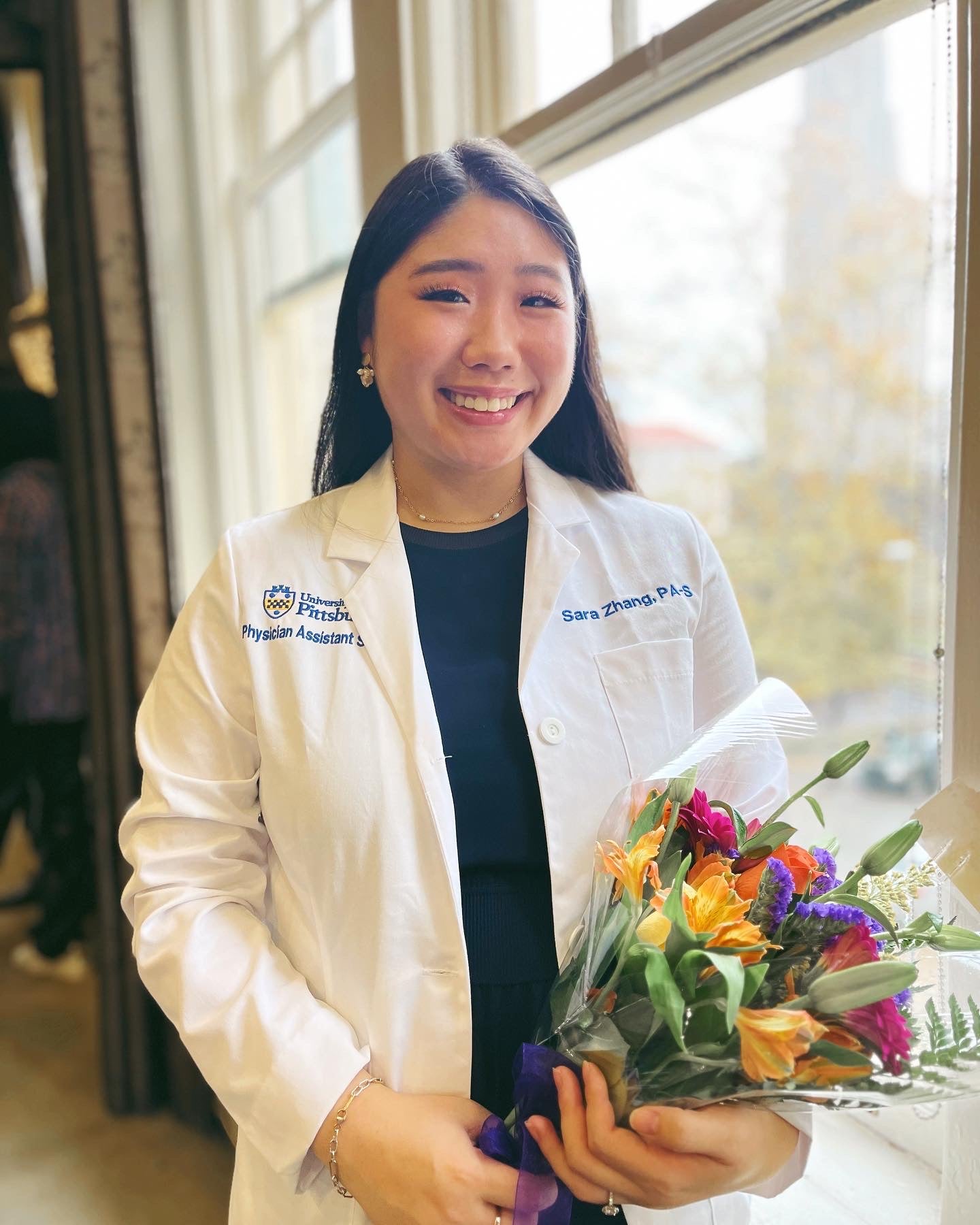 Woman with dark hair wearing a white lab coat over a dark dress and holding flowers.