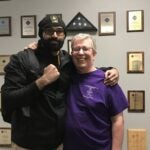 A man with a dark beard wearing a black beanie, a black zip-up and a backpack stands with his arm around a man with gray hair and black glasses who is wearing a purple t-shirt.