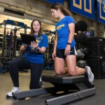 A woman with brown hair wearing a blue t shirt with a yellow Pitt panther on it, black leggings and white sneakers crouches on one knee while pointing to an iPad in her hand while speaking to a woman with curly brown hair pulled back who is wearing a blue shirt that says "Panthers Soccer" on it, black shorts and white sneakers who has her knees on a piece of Sports Science equipment.