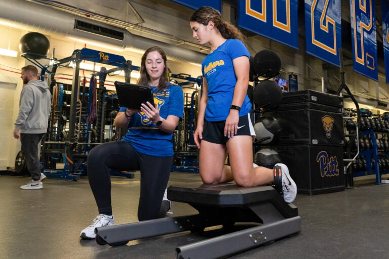 A woman with brown hair wearing a blue t shirt with a yellow Pitt panther on it, black leggings and white sneakers crouches on one knee while pointing to an iPad in her hand while speaking to a woman with curly brown hair pulled back who is wearing a blue shirt that says "Panthers Soccer" on it, black shorts and white sneakers who has her knees on a piece of Sports Science equipment.