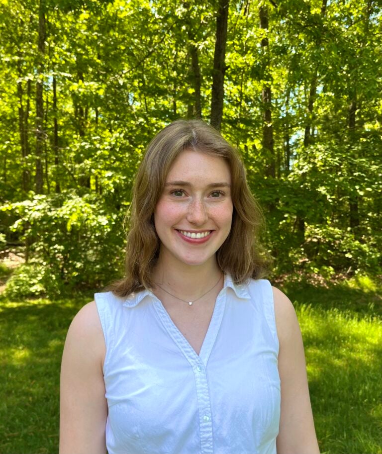 A woman with light brown hair wearing a sleeveless button down shirt with a collar and s silver necklace while standing outside.