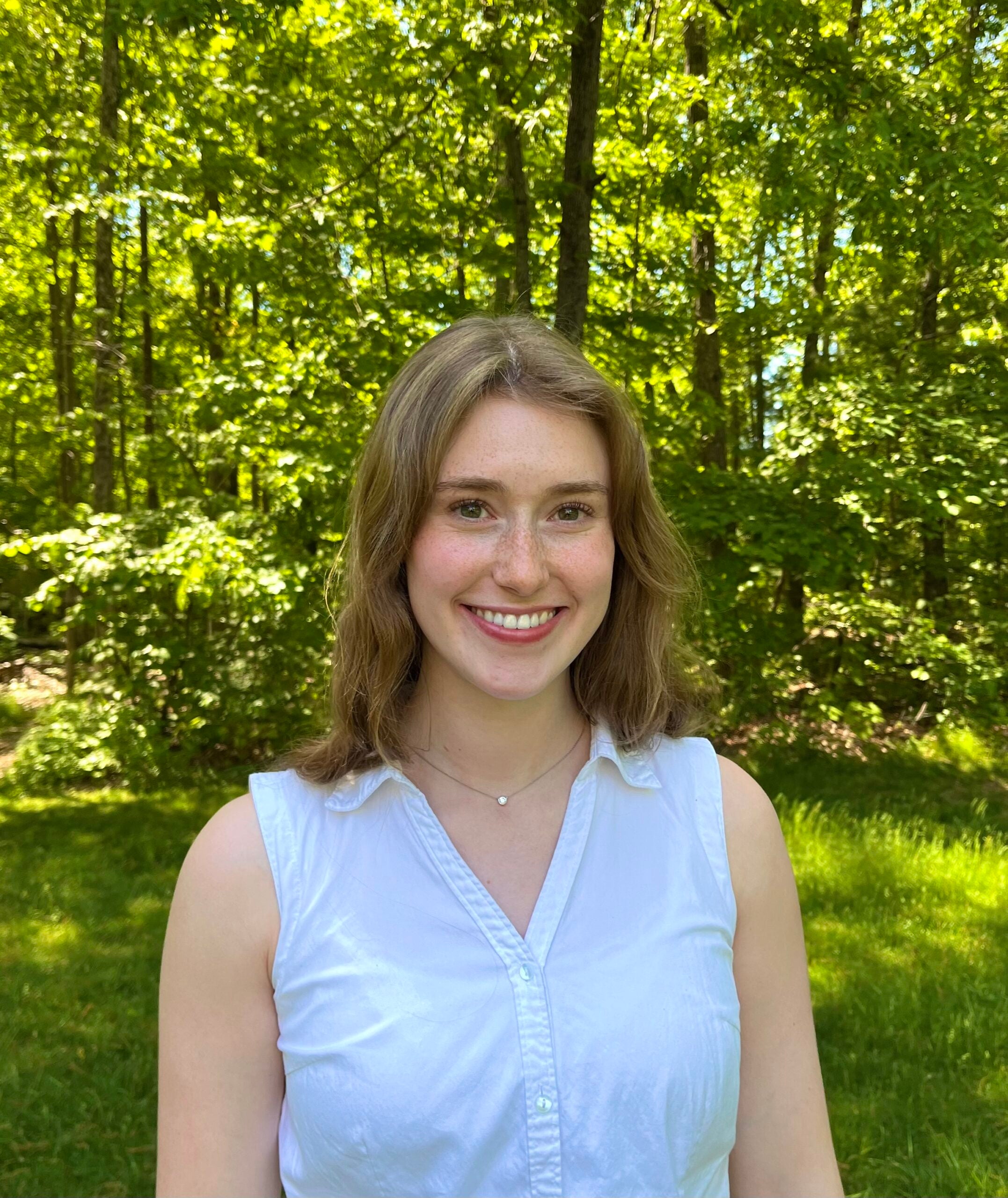 A woman with light brown hair wearing a sleeveless button down shirt with a collar and s silver necklace while standing outside.