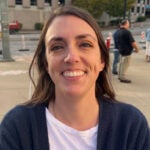 Woman with brown hair wearing a navy blue sweater over a white shirt smiling outside.