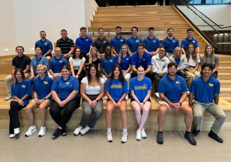 Three rows of students sitting and posing for a group photos. Students are wearing a variety of University of Pittsburgh themed shirts and attire.