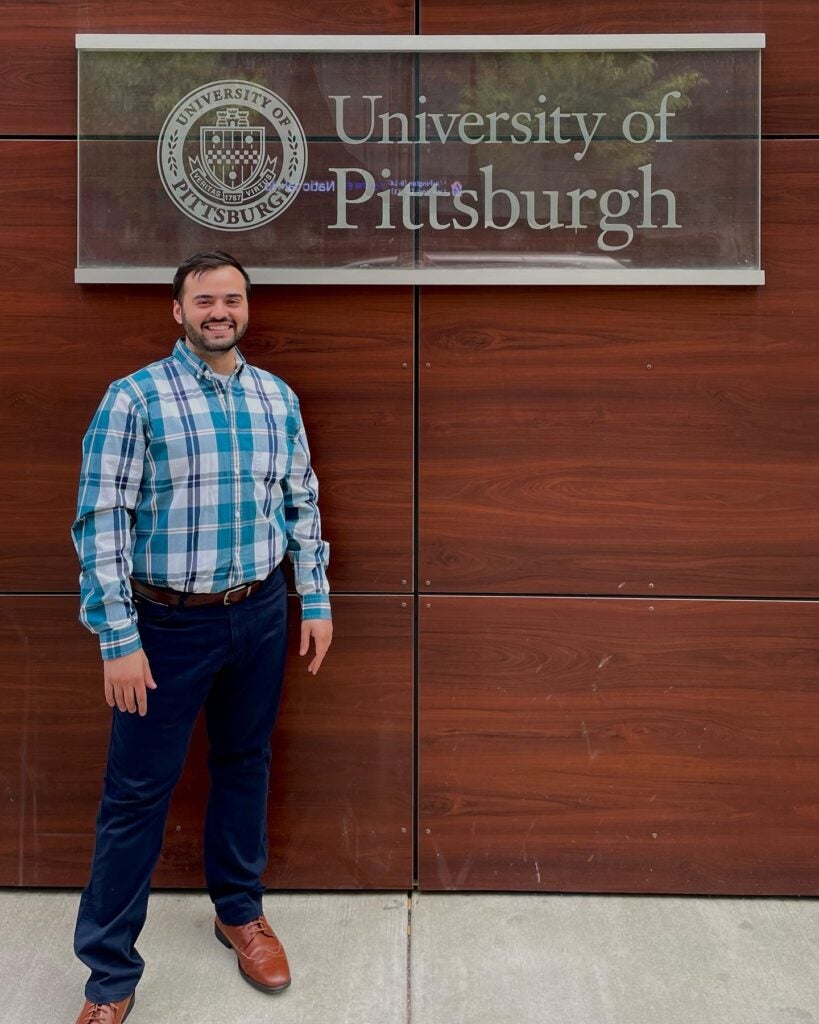 A man wearing a light and dark blue plaid collared shirt, navy pants and brown boots standing in front of a University of Pittsburgh sign.