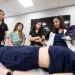 Three students wearing scrubs standing over a medical bed with a patient model. An instructor in a white coat shows the three students how to use the device in her hands which includes an ultrasound receiver and software on an iPad.
