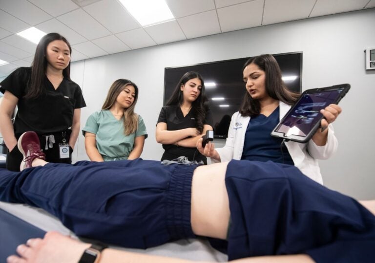 Three students wearing scrubs standing over a medical bed with a patient model. An instructor in a white coat shows the three students how to use the device in her hands which includes an ultrasound receiver and software on an iPad.