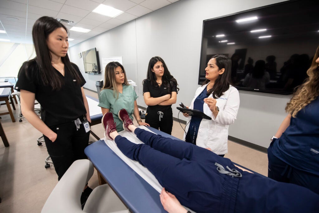 Three students in scrubs standing around a medical bed with a patient model on it. An instructor with a white coat on stands to the very right is holding an ultrasound receiver and an iPad.